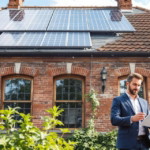Ein Mann in Anzug steht vor einem Backsteinhaus mit vier großen Fenstern und Solarpanels auf dem Dach, Symbol einer Energieeffiziente Altbau Modernisierung.