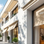 Helles Reihenhaus mit ausgefahrenen Markisen an den Fenstern als effektiver Hitzeschutz im Sommer in einer ruhigen Wohnstraße mit grünen Bäumen und modernem Mobiliar innen sichtbar durch das Fenster.